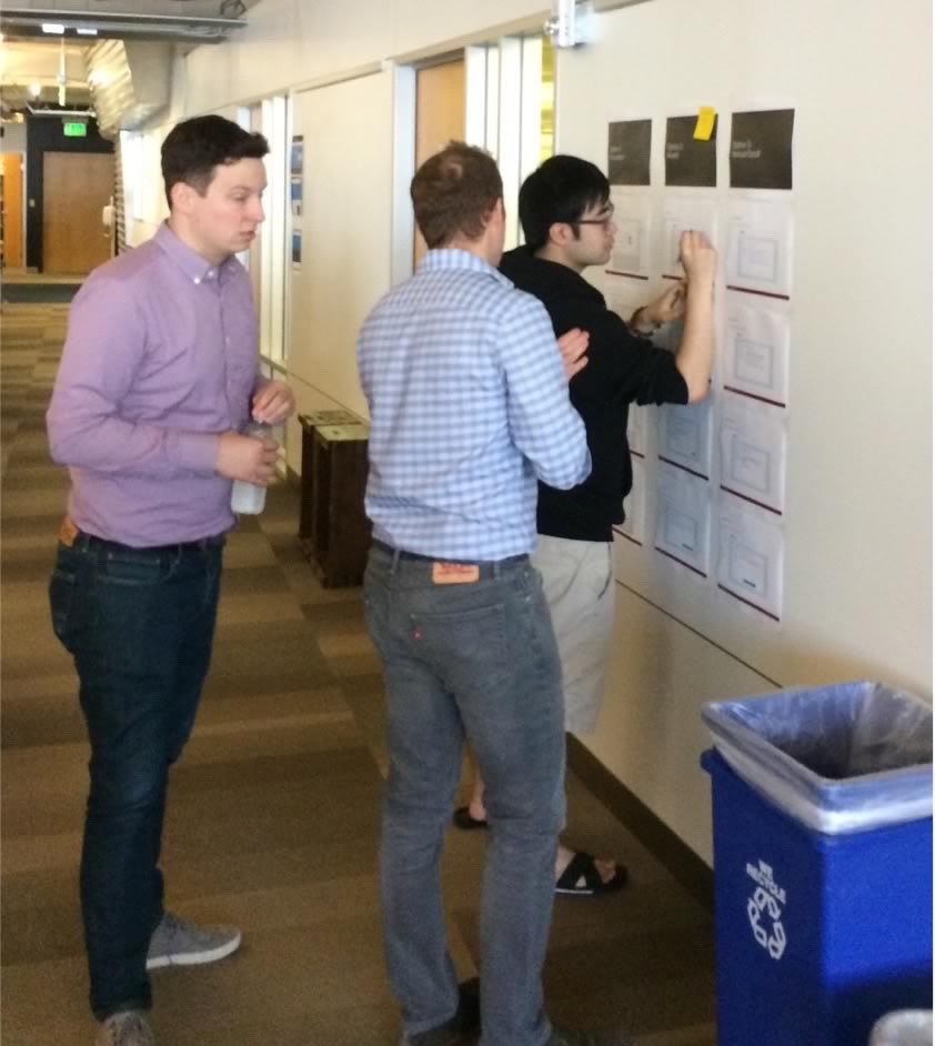 Three people at a wall of printed concept cards in a Docker hallway, one annotating a card